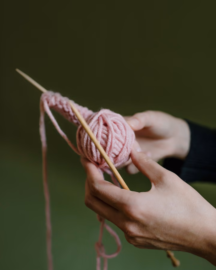 Händer som stickar med rosa ullgarn på två stickor, mot en grön bakgrund.